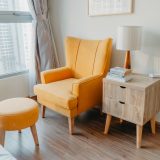 yellow armchair and stool beside wooden nightstand by the wall near glass window and bed